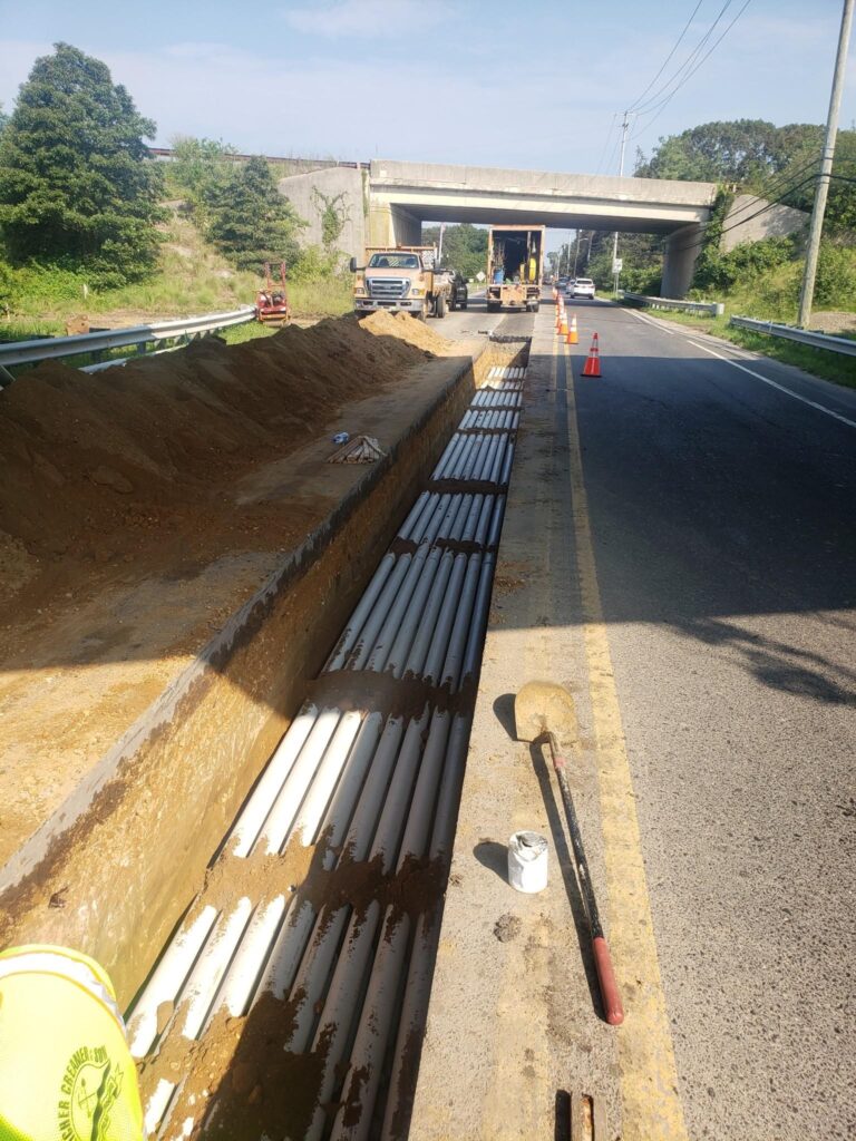 A trench is being dug alongside a roadway for the installation of multiple underground utility conduit